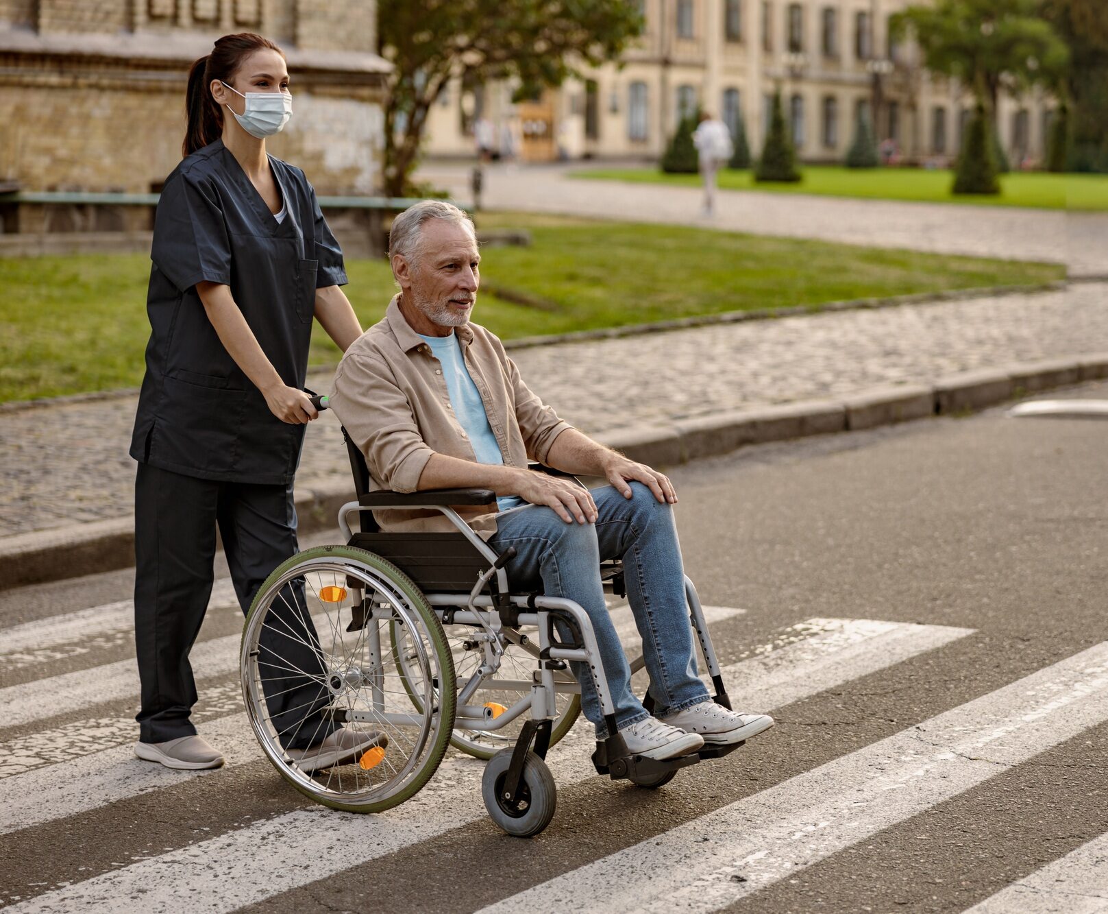 Full length shot of young nurse in protective face mask taking care of mature disabled man in
